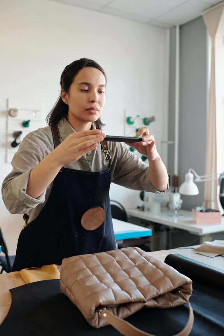 Young craftswoman in apron taking photo of handbag on smartphone